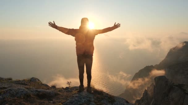 Man stands on the edge of a high mountain above the sea, spreads his arms to the sides and waves, covering his body with the shining sun, a close view. A bearded tourist stands near a cliff at sunset. - Footage, Video