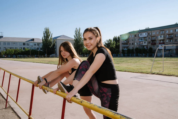 Two young athletic women in black tracksuits train at the stadium. Stadium on background. girls are doing exercises, stretching, yoga. Sport and healthy lifestyle. - Photo, Image