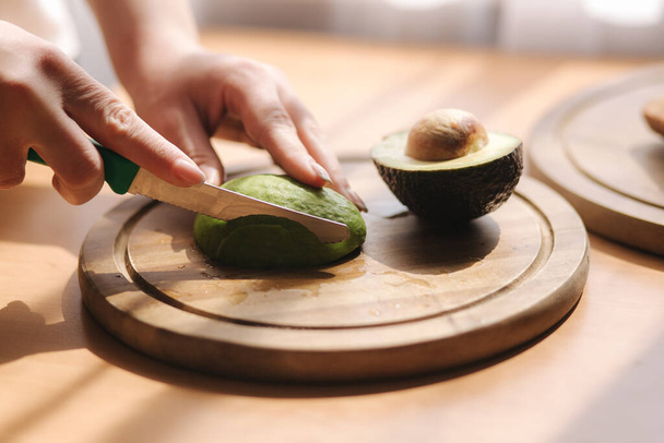 Close up of woman slicing avocado on wooden board at home. Vegetarian food concept - Foto, Bild