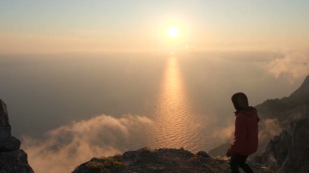 Sportos férfi turista fut fel a szélén egy magas hegy, és csodálja a lenyűgöző gyönyörű táj naplemente a tenger felett, és a füst a felhők. Aktív élet és túrázás a hegyekben és sziklákban. - Felvétel, videó