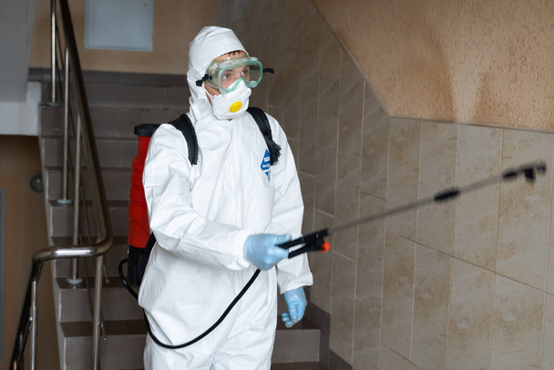 UKRAINE, KYIV - May 20, 2020: Man in a white protective suit and mask is sanitizing interior surfaces inside buildings while the coronavirus epidemic for infection prevention and control of epidemic. - Fotó, kép