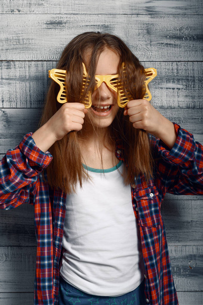 Funny little girl in headphones and star glasses in studio. Children and gadget, kid isolated on wooden background, child emotion, schoolgirl photo session - Photo, Image