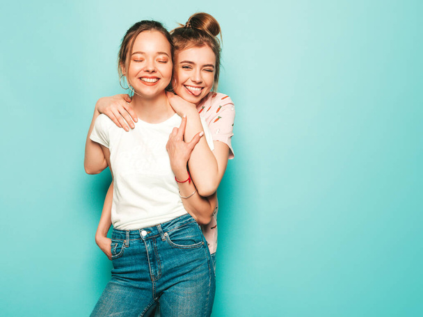 Two young beautiful blond smiling hipster girls in trendy summer hipster jeans clothes. Sexy carefree women posing near blue wall. Trendy and positive models having fun - Foto, Imagen