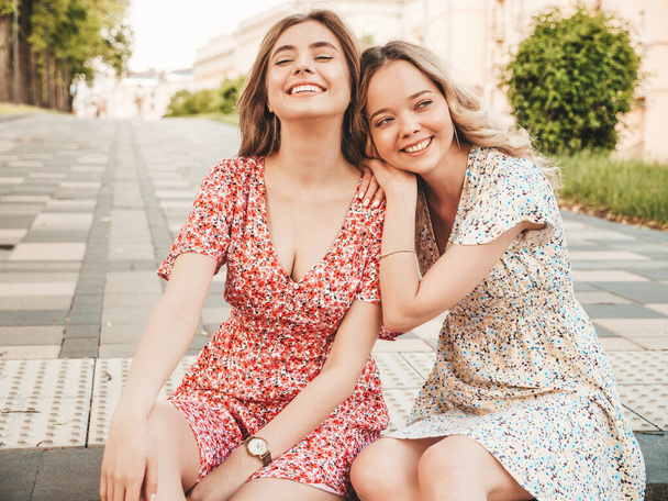 Two young beautiful smiling hipster girls in trendy summer sundress.Sexy carefree women sitting on the street background. Positive models having fun and hugging.They going crazy - Foto, Imagen