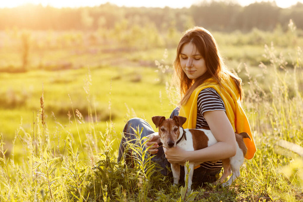 Красивая молодая девушка сидит на траве и обнимает свою собаку. Девушка и её питомец отдыхают после прогулки на природе. Друг, Дружба, Забота. Пространство для текста
 - Фото, изображение
