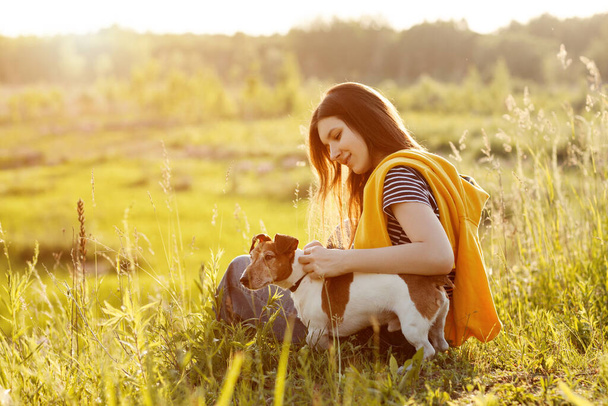 Ein schönes junges Mädchen sitzt auf dem Gras und umarmt ihren Hund. Ein Mädchen und ihr Haustier ruhen sich nach einem Spaziergang in der Natur aus. Freund, Freundschaft, Fürsorge. Raum für Text - Foto, Bild