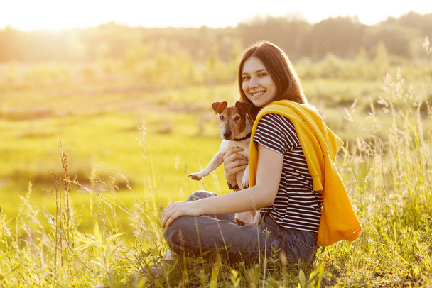 Ein schönes junges Mädchen mit langen dunklen Haaren umarmt einen weißen Jack Russell Terrier. Ein Mädchen sitzt mit ihrem Hund im Gras in der Sonne. - Foto, Bild