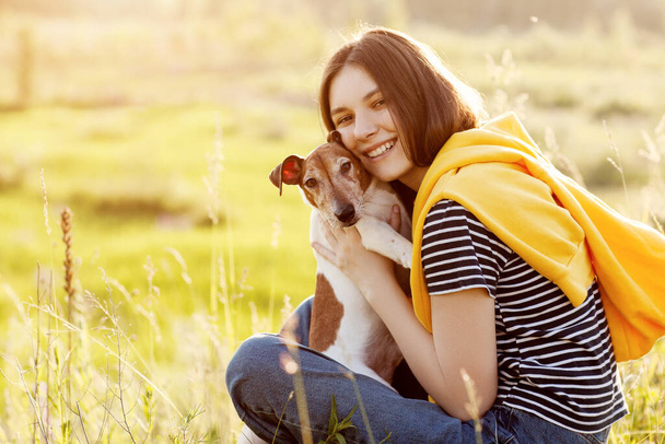 A charming girl is sitting on the grass, smiling and hugging her dog. A girl and her pet are relaxing in the fresh air. Friend, friendship, care - Photo, Image
