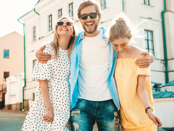 Group of young three stylish friends posing in the street. Fashion man and two cute girls dressed in casual summer clothes. Smiling models having fun in sunglasses.Cheerful women and guy at susnet - Фото, изображение