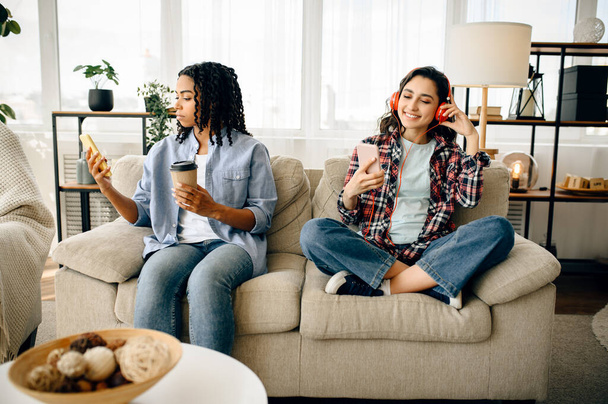 Two women in headphones leisures on sofa. Pretty girlfriends in earphones relax in the room, sound lovers resting on couch, female person using mobile phone - Foto, immagini