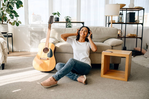 Mosolygó nő fülhallgatóban otthon, gitár a háttérben. Csinos hölgy hangszer pihenni a szobában, női zene szerető pihen - Fotó, kép