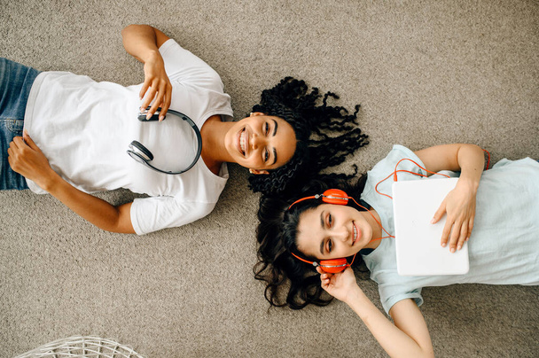 Δύο γυναίκες με ακουστικά απολαμβάνουν να ακούνε μουσική ξαπλωμένη στο πάτωμα. Όμορφες κοπέλες με ακουστικά χαλαρώνουν στο δωμάτιο, οι λάτρεις του ήχου αναπαύονται στον καναπέ. - Φωτογραφία, εικόνα
