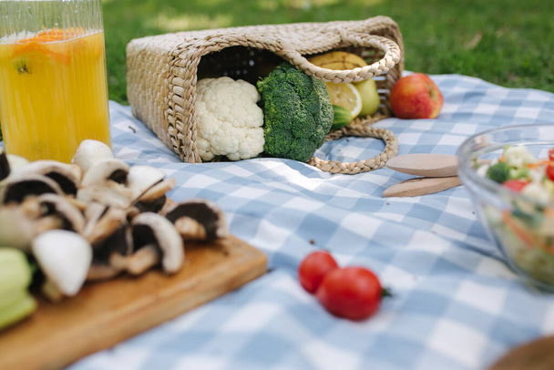 Cesta de picnic con sándwiches veganos saludables en manta a cuadros azules en el parque. Frutas frescas, verduras y zumo de naranja. Concepto de picnic vegano
 - Foto, Imagen