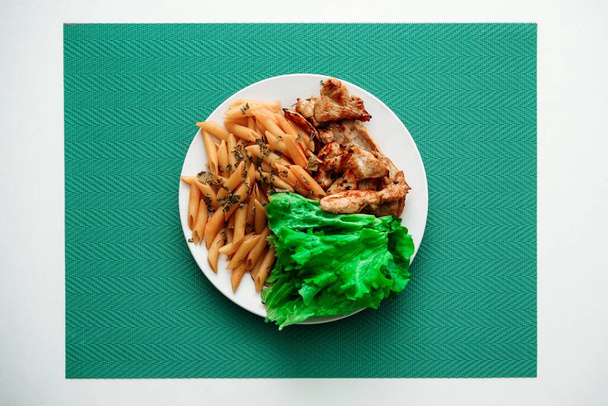 Un plato con pasta de penne cocida, pollo asado y lechuga sobre un tejido de color turquesa
 - Foto, imagen