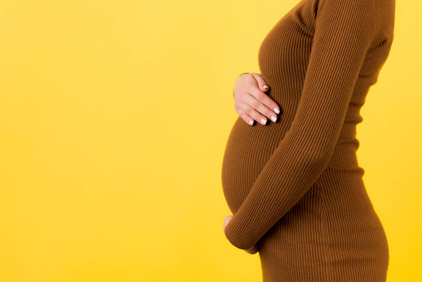 Καλλιεργημένη εικόνα της εγκύου που αγκαλιάζει την κοιλιά της. Η μητέρα φοράει καφέ φόρεμα σε κίτρινο φόντο. Ο μελλοντικός γονιός περιμένει το παιδί της. Αντιγραφή χώρου. - Φωτογραφία, εικόνα