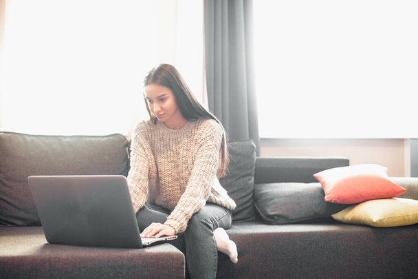 Young woman with modern laptop sitting on sofa at home. Happy girl browsing through the internet during free time..  - Foto, Imagen