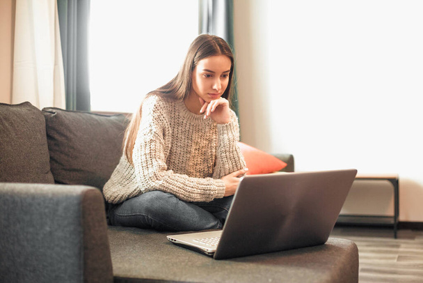 Young woman with modern laptop sitting on sofa at home. Happy girl browsing through the internet during free time..  - Foto, Bild