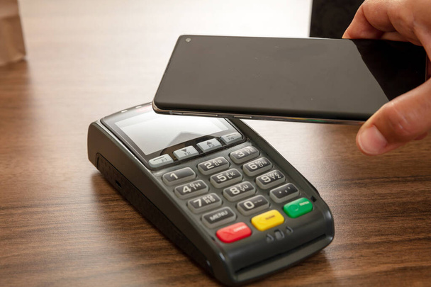 Mobile payment, smart phone nfc, near field communication wireless technology. Customer hand holding a smartphone close to a POS terminal, closeup view - Photo, Image