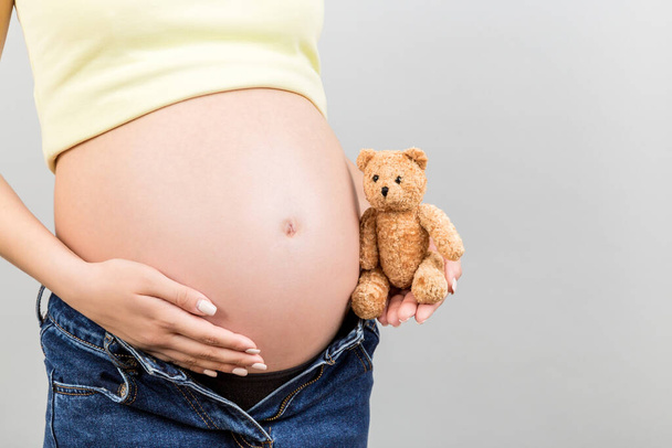 Close up of pregnant woman in unzipped jeans embracing teddy bear near her belly at colorful background with copy space. Expecting a baby concept. - Foto, Imagem