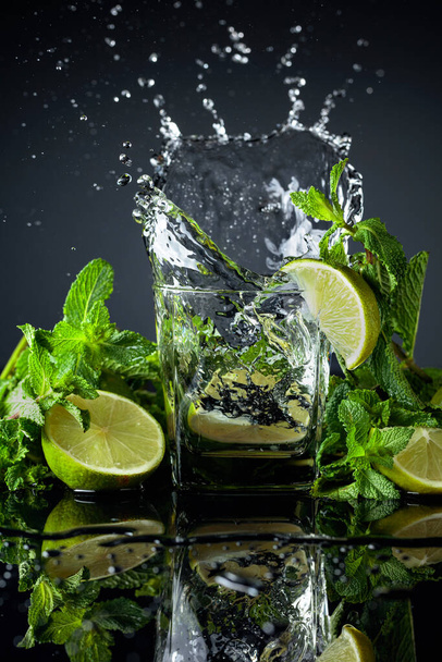 Cocktail com limas e hortelã em um fundo reflexivo preto. Fatia de limão cair em vidro com bebida
. - Foto, Imagem