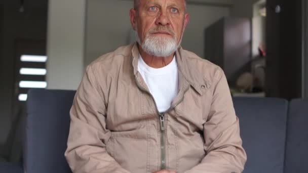 A man with a gray beard sits on a sofa with a sad face. Dismissed worker worries about future, unemployment and economic crisis concept - Materiał filmowy, wideo