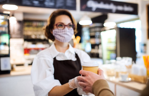 Young woman with face mask working indoors in coffee shop, serving customer. - 写真・画像
