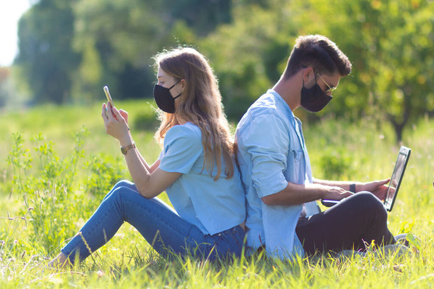 公園に座っている2人の若者がノートパソコンとスマートフォンをフェイスマスクで使用しています-コロナウイルスの時代の学生 - 写真・画像