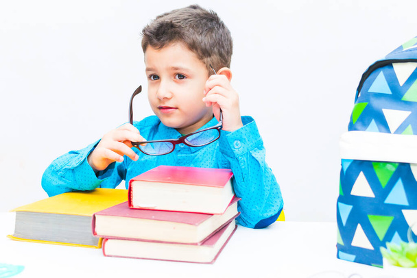 Een kleine donkerharige kleine schattige jongen in een blauw shirt zit aan een tafel met boeken en zet een bril op. - Foto, afbeelding