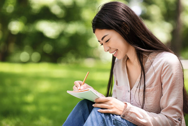 Молода азіатська студентка сидить на газоні в парку і пише нотатки
 - Фото, зображення