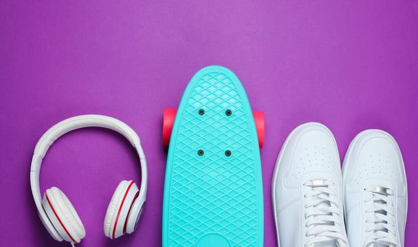 Hipsterská výbava. Skateboard se sluchátky a tenisky na fialovém pozadí. Kreativní módní minimalismus. Módní staromódní styl. Minimální letní zábava. Hudební koncept - Fotografie, Obrázek