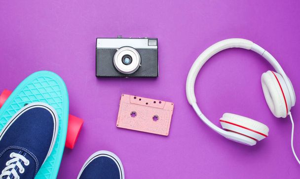 Hipster fashion look. Skateboard, sneakers, audio cassette, retro camera, headphones on purple color background. 80s. Top view. Flat lay - Fotó, kép