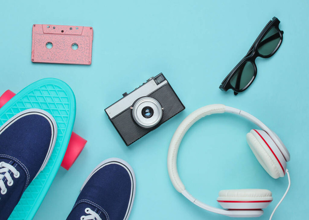 Hipster outfit. Skateboard with headphones, audio cassette, retro camera and sneakers on blue background. Creative fashion minimalism. Minimal summer fun. Top view - Photo, Image