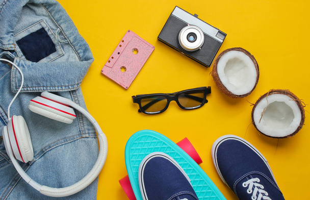 Hipster outfit. Skateboard with headphones, jeans jacket, audio cassette, retro camera, coconut and sneakers on yellow background. Creative fashion minimalism. Minimal summer fun. 80s. Top view - Photo, Image