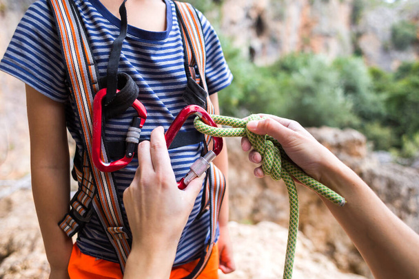 インストラクターは子供にクライミング機器を使用することを教え、少年は岩に登る準備をしており、女性は子供に、安全システムにロープを結ぶ、と主張するためにキャビナーを使用する方法を示しています。. - 写真・画像