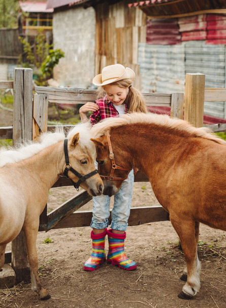 little blond hair girl in cowboy hat and boots pink plaid shirt with pony little horse - Valokuva, kuva