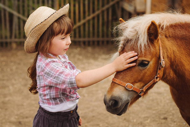 μικρή ευτυχισμένη όμορφη κοπέλα με καστανά μαλλιά σε καουμπόικο καπέλο και μπότες σε ροζ καρό πουκάμισο με μπεζ πόνυ μικρό άλογο - Φωτογραφία, εικόνα