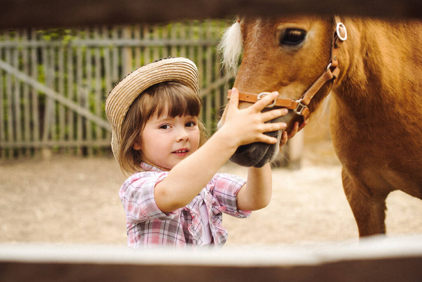 little happy beautiful girl with brown hair in cowboy hat and boots in pink plaid shirt with beige pony little horse - Fotó, kép