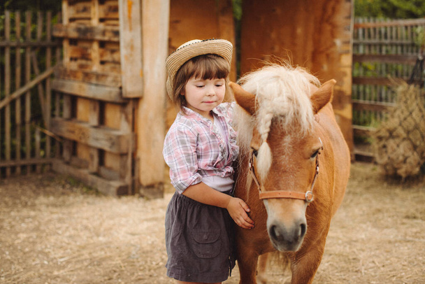 klein gelukkig mooi meisje met bruin haar in cowboy hoed en laarzen in roze geruite shirt met beige pony klein paard - Foto, afbeelding