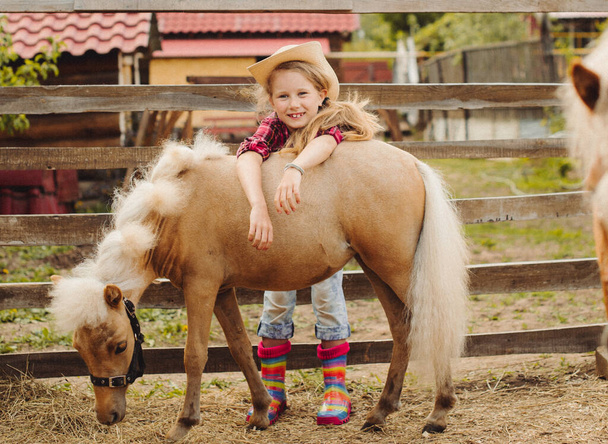 little blond hair girl in cowboy hat and boots pink plaid shirt with pony little horse - Fotoğraf, Görsel