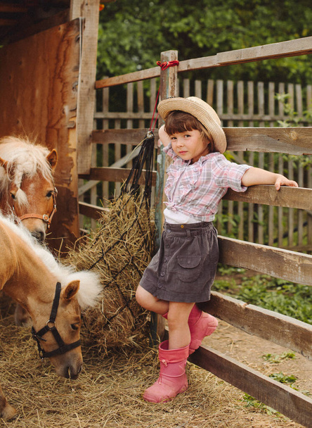 little happy beautiful girl with brown hair in cowboy hat and boots in pink plaid shirt with beige pony little horse - Foto, afbeelding
