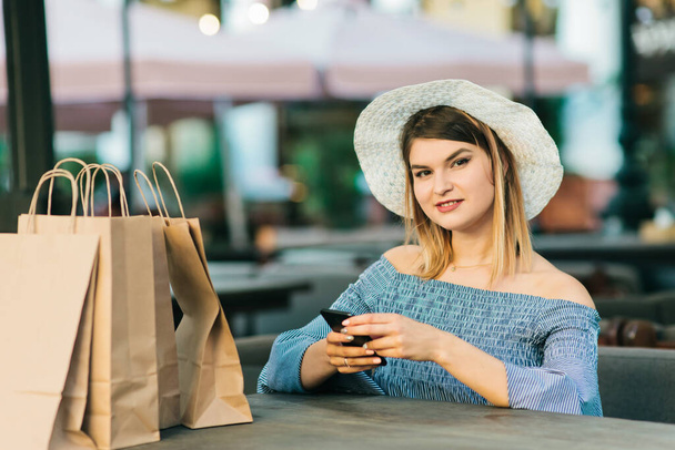 スタイリッシュなドレスと帽子の魅力的なお店の女性は屋外カフェのテーブルに座って、スマートフォンを使用しています - 写真・画像