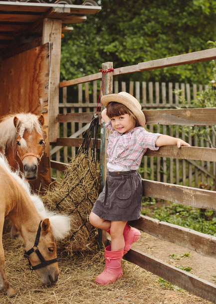 little happy beautiful girl with brown hair in cowboy hat and boots in pink plaid shirt with beige pony little horse - Fotografie, Obrázek