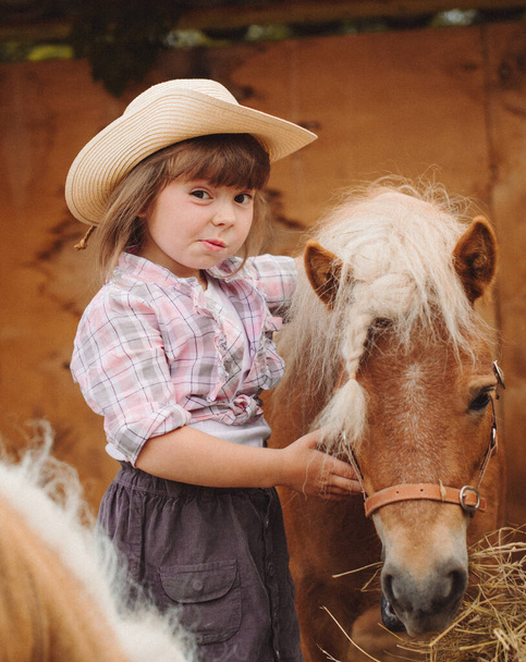 little happy beautiful girl with brown hair in cowboy hat and boots in pink plaid shirt with beige pony little horse - Foto, Imagen