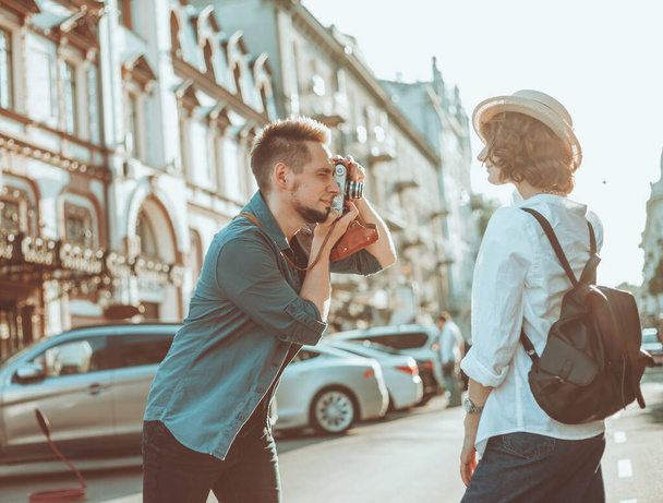 Young cheerful couple of hipsters are photographed on a retro camera while walking around the city. Male photographer photographs a stylishly dressed young woman - Fotografie, Obrázek