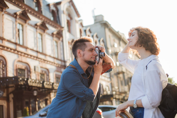 Молодые веселые хипстеры фотографируются на ретро-камеру во время прогулки по городу. Мужчина-фотограф фотографирует стильно одетую молодую женщину - Фото, изображение