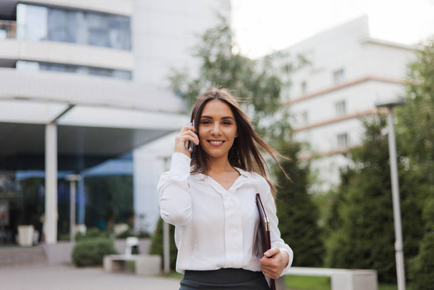 Bizneswoman. Młoda atrakcyjna menadżerka w spódnicy i bluzce rozmawiająca przez telefon w tle miejskiego krajobrazu. Wszystko w biznesie - Zdjęcie, obraz
