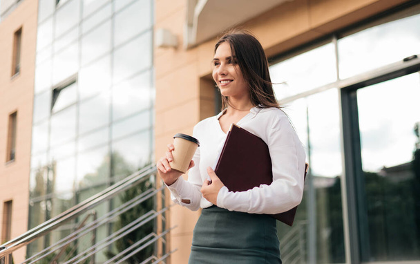 Joven mujer de negocios profesional con una blusa y una falda saliendo del centro de negocios y bajando las escaleras mientras bebe café al aire libre - Foto, Imagen