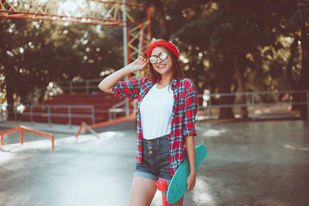 Jeugdsport. Jonge aantrekkelijke hippe vrouw met een rood geruit shirt en spijkerbroek met een skateboard in een skatepark. Zomertijd - Foto, afbeelding
