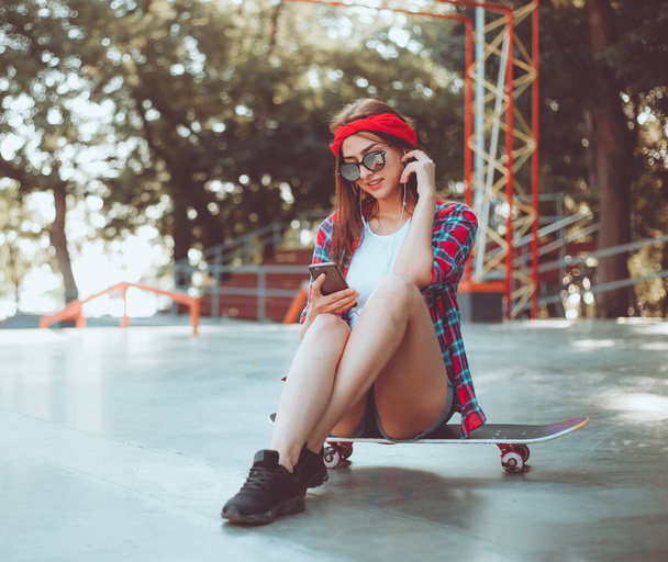 Fiatal elegáns nő öltözött ifjúsági ruhák ül egy gördeszkán, és zenét hallgat a fejhallgató egy gördeszka. Nyári időszak - Fotó, kép