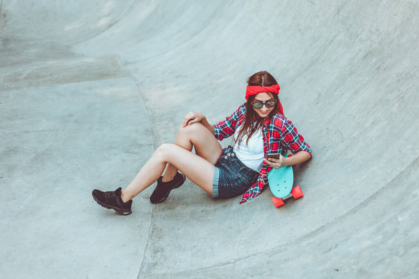 Youth pastime. Young hipster woman resting with a skateboard at a skatepark - Фото, изображение